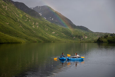 Bride and groom walk hand in hand along a remote Alaskan beach, surrounded by misty mountains and a moody overcast sky. The groom wears a plaid shirt and jeans, while the bride’s wedding dress flows behind her, capturing the quiet beauty of an intimate Alaska elopement.