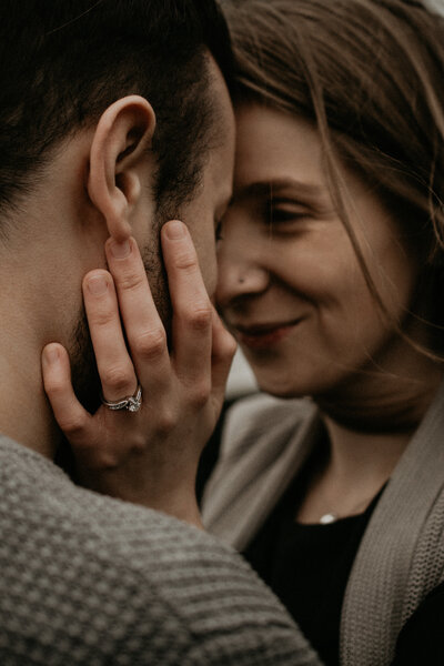 close up of a couple touching their foreheads together. The angle is over the guy's shoulder, so you can see part of the girls face as she looks at him. Her hand is resting on his cheek. The focus is on her engagement ring.