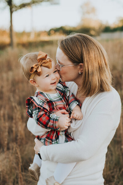 Mom kisses baby girl in a field during fall family session in Raleigh NC