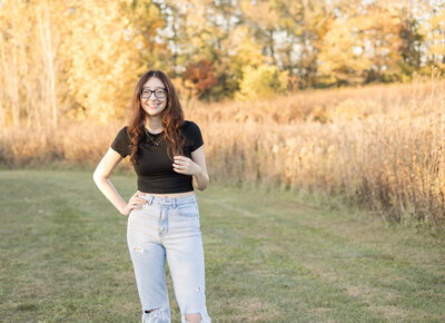 high school senior female with long dark hair, standing in grass with trees behind her, wearing blue jeans, and a black short sleeved shirt