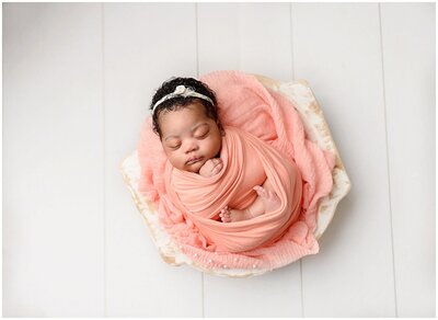 A newborn baby girl with a full head of dark curly hair in peach wrap laying in the bowl with feet out.