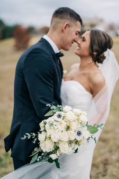 man in black suit leaning in to woman in white dress and veil holding a white bouquet with greenery