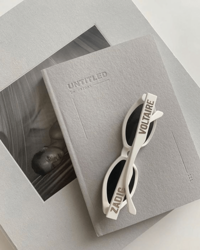 A stylish flat lay featuring Voltaire-branded white sunglasses placed on top of a grey 'Untitled' journal, with a monochrome photograph underneath, creating a minimalist and artistic aesthetic.