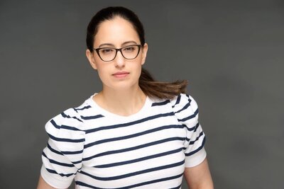 A woman with brunette hair wears a white and black striped shirt with black glasses