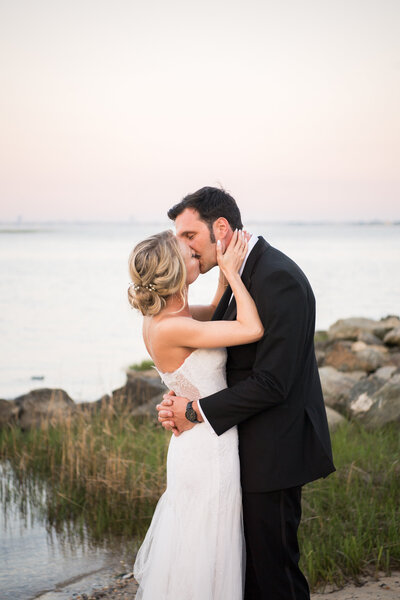 Bride and groom kissing as the sun is just setting creating a gentle hue of pinks in the sky with water and marsh in the backround
