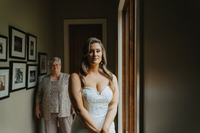 Bride in wedding dress, looking to camera as mom watches from being. 