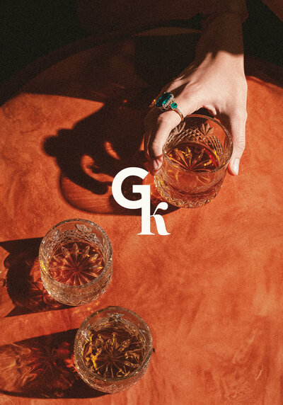 Three crystal cocktail glasses casting shadows on red velvet, overlaid with the GK submark logo, showcasing Gary & Kim’s coaching brand identity.
