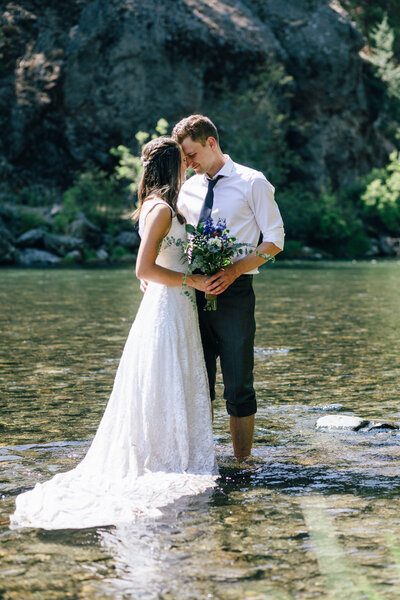 A couple in their wedding clothes stands in the Naches River at Whistlin' Jacks for their Mt. Rainier National Park elopement in Washington.