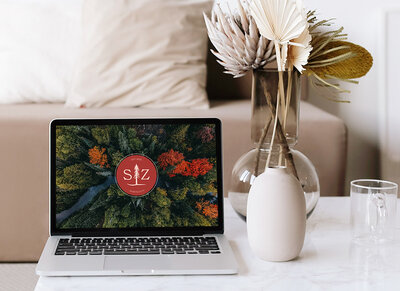 A white table with two vases and a laptop is visible in the foreground. The laptop is open to Sheri Zanganeh Therapy's submark logo, surrounded by an image of lush green, orange, and red trees. A beige couch and two pillows is visible in the background.