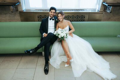 Newlywed couple sitting on a modern couch, smiling and embracing, with the bride holding her bouquet and the groom in a classic tuxedo.