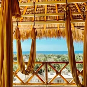 Golden aerial yoga hammocks hang inside a thatched palapa at Casa Calibra, overlooking the rooftops and lush palm trees of Puerto Escondido with the Pacific Ocean in the distance.