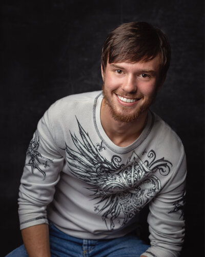 light-skinned smiling man in sitting in front of textured black background with left forearm on left lap and right hand hidden on the stool