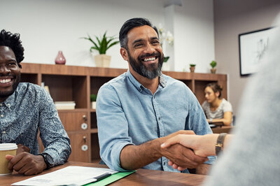 An Arab man shakes the hand of another colleague. He is wearing a blue shirt and has a beard.