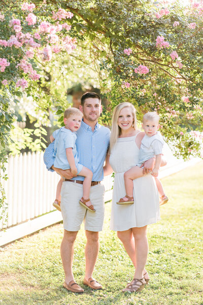 Family of four photograph in the spring in front of white picket fence and pink crape myrtle trees