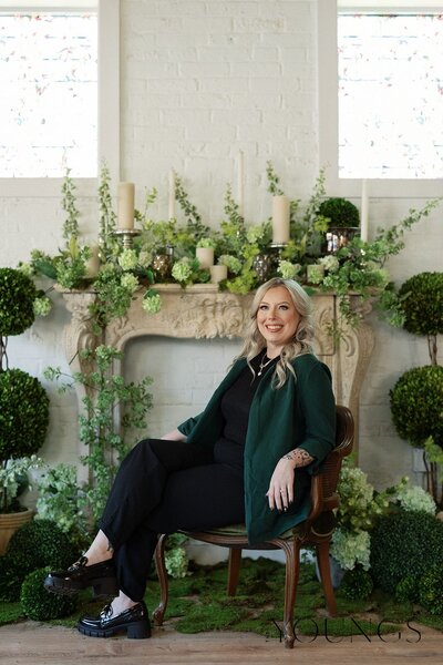 Jazmin, the co-owner, sitting sideways in a char, wearing a black outfit and green blazer. She's smiling. In the background, there's a mantel and a beautiful array or arranged greenery.