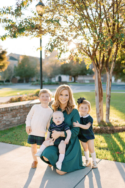 Mom of three, two boys and a girl, squats down with kids in front of her smiling. Sunlight streams through a tree behind them and there is a brick wall to their left in this downtown Statesboro photography spot.