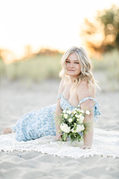 A woman lays on a blanket on the beach wearing a blue summer dress looking at the camera, photographed by Tambella Photography