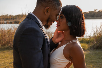 A romantic outdoor engagement photo at Bay Area Park in Houston, Texas, featuring a couple standing close to each others faces as the man gently holds the woman’s face, with warm golden-hour light reflecting off the lake in the background.