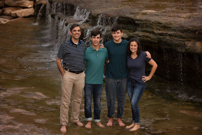 family-or-4-with-two-teen-boys-standing-by-waterfall-in-fort-worth-tx-wearing-blues-and-greens