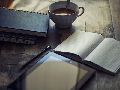 A milky cup of coffee sits on a light wood table amid a spiral-bound journal, and open lined notebook, a pencil, and a tablet.