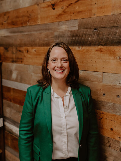 Kim McKenna wearing a green blazer and standing in front of a wood board wall