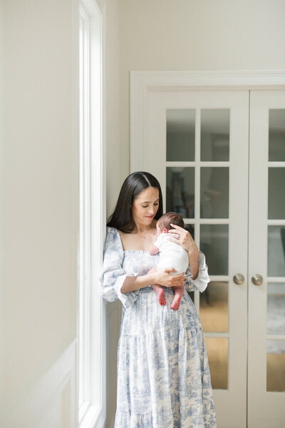 A woman wearing a blue dress leans against a window frame during her NJ newborn photos.