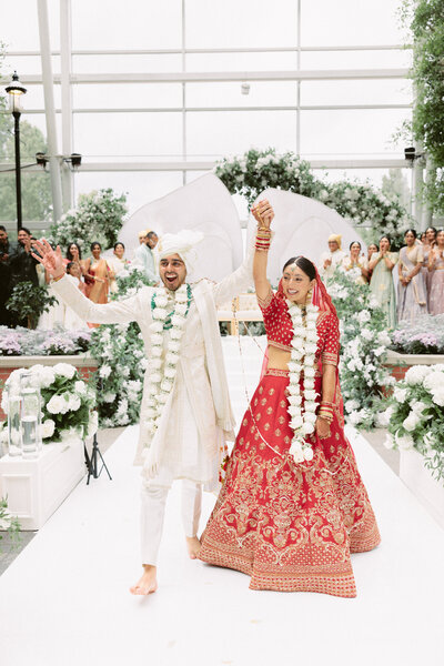 South Asian couple excitedly exits from their wedding ceremony in stunning lehengas and flower necklaces.