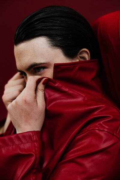Person wearing a deep red coat holding the collar up by their eyes with a deep red backdrop