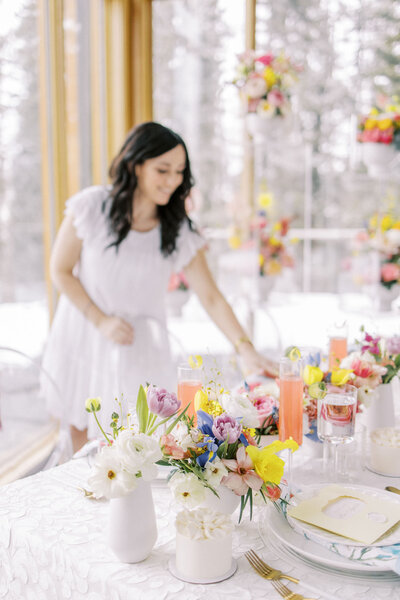 A woman in a white dress arranges items on a table set with floral arrangements, colorful drinks, and plates in a bright room with large windows, perfect for weddings in Banff Alberta.