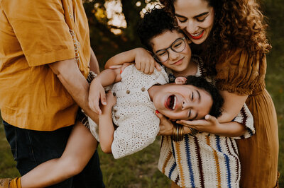 A little boy leaning sideways while him mom and his brother tickle him and laughs and giggles in fort worth, texas
