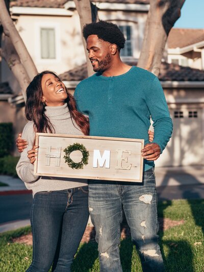 Black couple standing in front of their new home holding a sign that says home