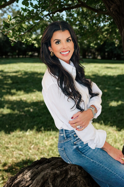 Professional outdoor headshot of a woman with long dark hair, wearing a white blouse and jeans, seated on a rock with greenery in the background in Little Rock, Arkansas.

