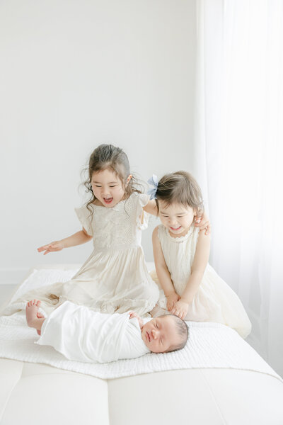 Newborn baby sleeps swaddled on a bed as his older sisters joyfully smile looking at him