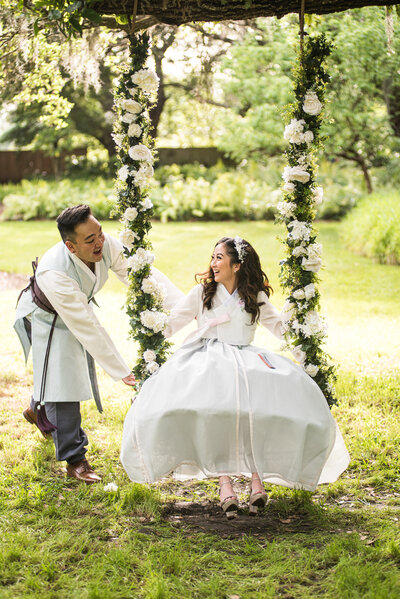 bride in traditional hanbok being pushed by groom on a flower draped swing