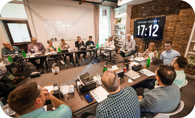 Large group seated around a U-shaped conference table, reviewing materials while a timer counting down is displayed on a screen