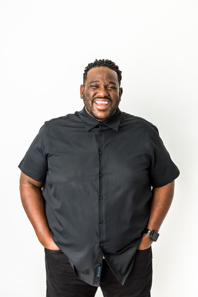 African American man in black shirt laughs at camera on a white backdrop.
