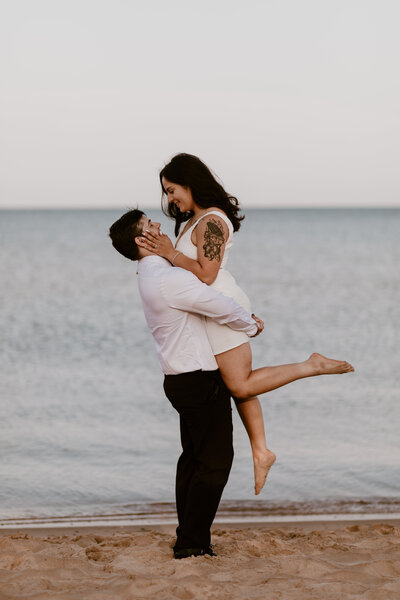 A couple embraces on the beach; the man in a white shirt lifts the woman in a white dress, showing joy. Calm sea and sandy shore set a romantic tone.