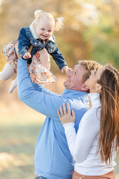 A family unit of 3, father is tossing up toddler into the air, mother is holding onto father watching the toddler, everyone is smiling.