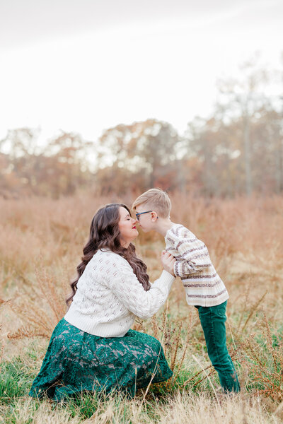 A mother in a green dress and white sweater nuzzles the nose of her toddler son in a field at sunset