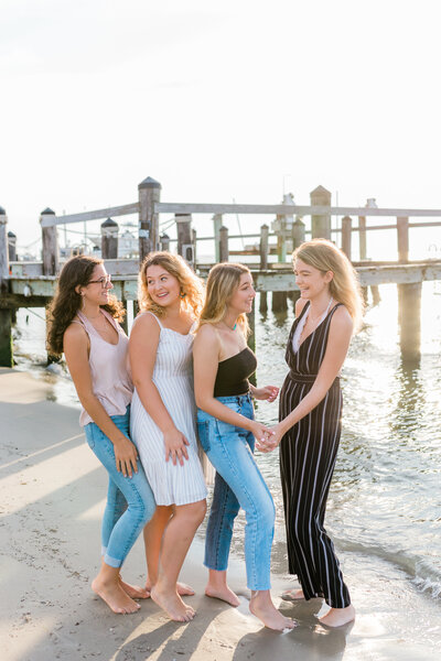 High school senior photographers. Four best friends posing for a photoshoot on the beach in Ocean City, New Jersey. They are holding hands and laughing. The sun is glowing on the water behind them.