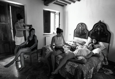 True documentary black and white wedding photo of the bridal prep behind the scenes on the morning of a destination wedding in Tuscany, Italy. Bride having her hair styled while her bridesmaids chill on the bed and one massages her pregnant sisters head
