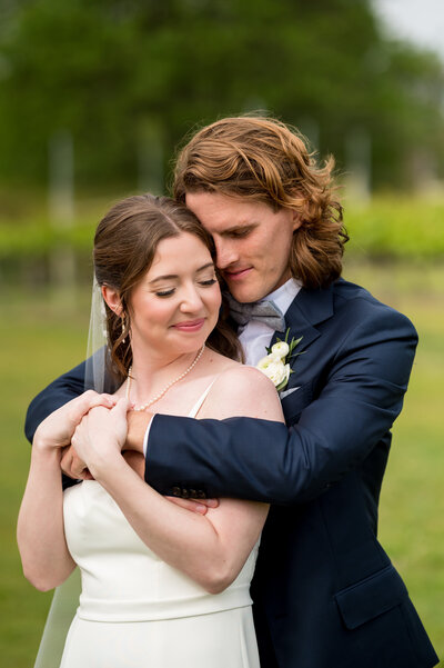 Bride and groom standing in the vineyard at Willow Creek Winery in Cape May NJ.  The bride is wearing pearls and a white gown and veil and the groom is wearing a custom blue suit with a gray bowtie. He is hugging her from behind and resting his head on hers with their eyes closed.