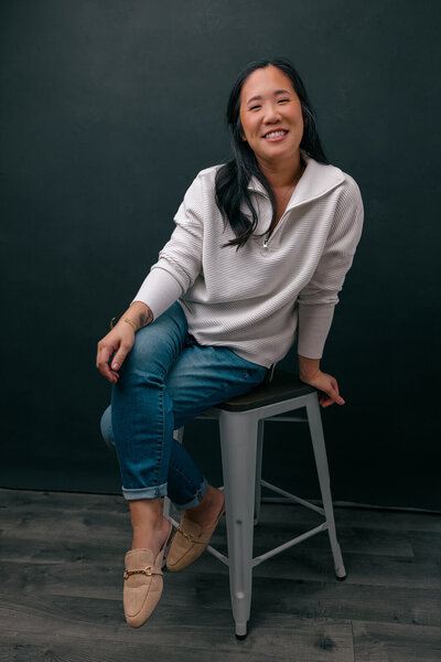 Asian woman wearing white sweater and jeans sitting on a stool, grinning at the camera