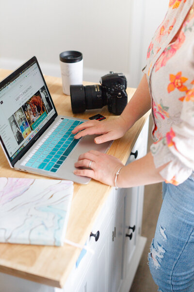 Kristin Palkoner's hands typing on a laptop, with Nikon camera, coffee cup and notebook on table.