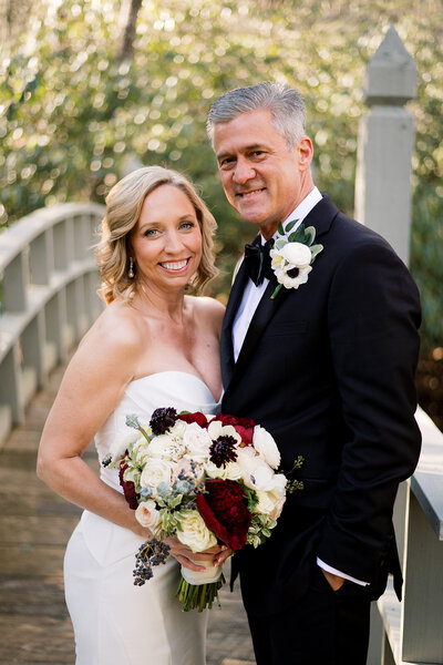 woman in white dress holding red and white flowers next to man in tux smiling at the camera