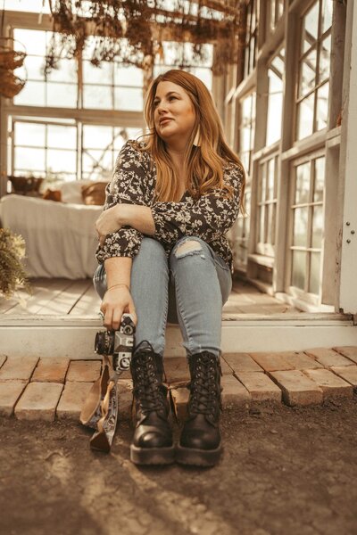 The journey with Gillian Oler's in a Dallas, TX greenhouse. The  professional in family photography sits amidst nature, framed by elegant white doors, showcasing her passion with a black camera in hand. Dressed in a stylish ensemble of blue jeans, a floral blouse, and black boots, Gillian exudes creativity and flair.