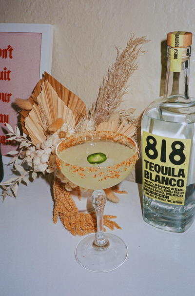 A close-up of a spicy margarita cocktail garnished with a jalapeño slice. The cocktail is in a stemmed glass with a salted rim. A bottle of 818 Tequila sits behind the glass, and a dried floral arrangement is in the background.