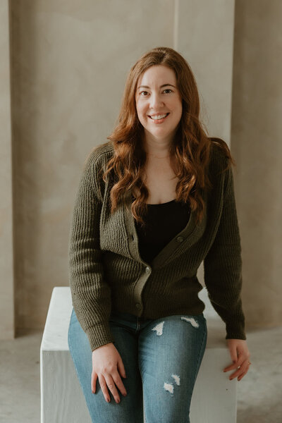 Virtually Robyn, a woman and virtual assistant, with long brown hair and a green sweater smiles at the camera while leaning against a box. 