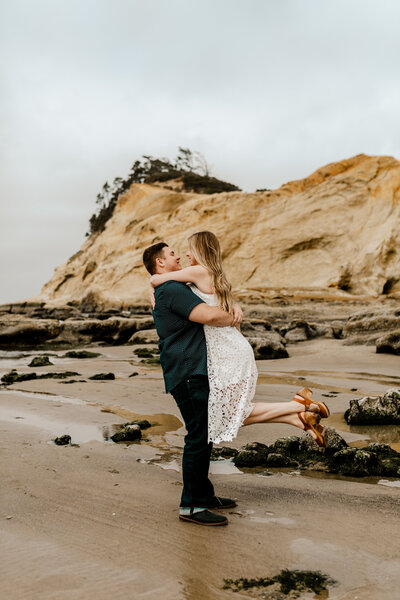 Couple embracing on the cliffs of Cape Kiwanda at sunset, sandstone bluffs glowing golden above the waves.