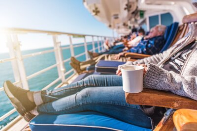 People relax in chaise lounge chairs on a cruise deck while looking out across the water and enjoying cups of coffee.